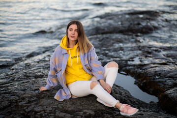 a girl sits on a rocky surface in a gray coat and a yellow hoodie and white torn jeans against the backdrop of nature