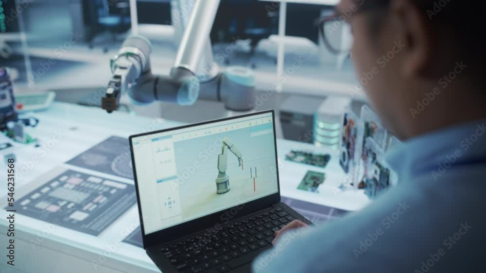 Poster Over the Shoulder Shot of a Robotics Engineer Using a Laptop Computer with a Software Operating the Robotic Arm. Robot Hand Holding a Microchip. Automation Startup Research and Development Office.