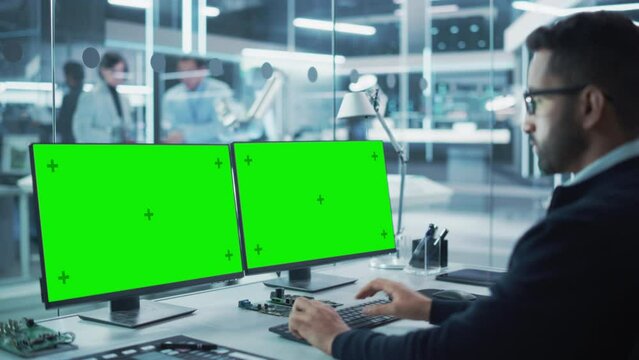 Software Developer Working on a Desktop Computer with Two Green Screen Chromakey Display Screens in a Factory Facility. Modern Technological Research and Development Center.
