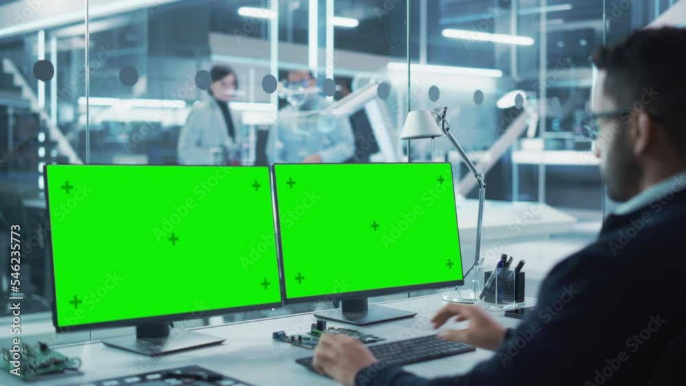 Poster Robotics Engineer Working on a Desktop Computer with Two Green Screen Chromakey Display Screens in a Startup Factory. High Tech Technological Research and Development Company.