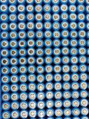 A close-up image of a grid of cylindrical lithium-ion batteries. The batteries are arranged in rows and columns, with their blue plastic casings and silver metallic tops clearly visible.