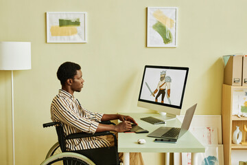 Side view of young male designer in wheelchair creating new graphic pictures while sitting by desk and looking at image of warrior on screen