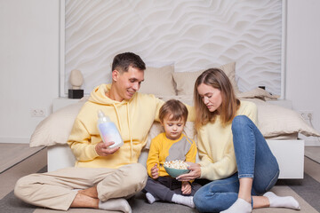 Cute adorable family sitting by the bed, dad holding bottle, mom and son eating popcorn