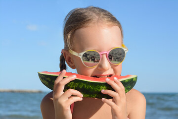 Cute little girl with sunglasses eating juicy watermelon on beach, closeup