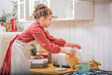 A little girl in the kitchen in an apron cooks something in a saucepan on a gas stove.