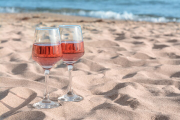 Glasses of tasty rose wine on sand near sea, space for text