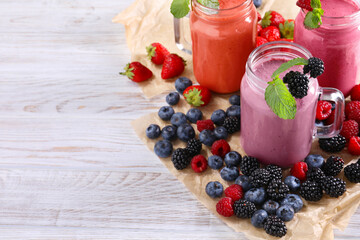 Mason jars of different berry smoothies and fresh ingredients on white wooden table. Space for text