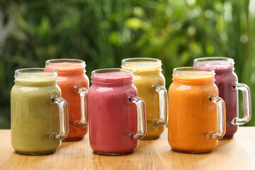 Many different delicious smoothies in mason jars on wooden table against blurred background