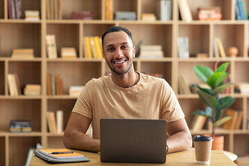 Smiling arab man sitting at desk, working on pc laptop indoors in living room, copy space. Distant job concept