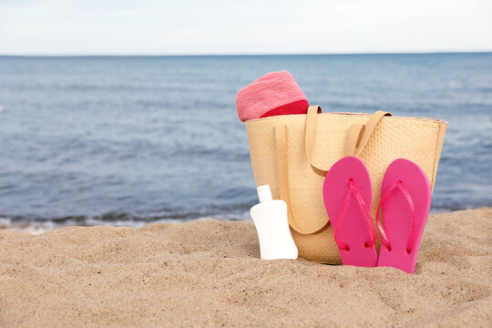 Summer bag with slippers, beach towel and sunscreen on sand near sea, space for text