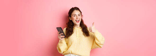 Portrait of attractive glamour girl showing thumbs up after using app on smartphone, online...