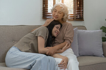 Loving adult daughter hugging older mother on couch at home, family enjoying tender moment together, young woman and mature mum or grandmother looking at each other, two generations