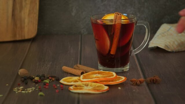 Woman's hand placing glass with mulled wine on wooden table. red hot wine alcohol drink with apple and orange fruit poured in glass, decorated with cinnamon stich and spice, slow 4k video footage