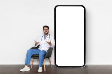 Telemedicine Concept. Handsome Male Doctor Sitting In Chair Near Big Blank Smartphone