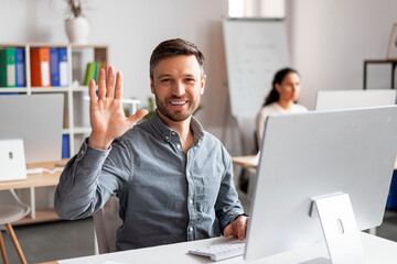 Smiling attractive millennial caucasian man manager working at computer, waving hand at camera - obrazy, fototapety, plakaty