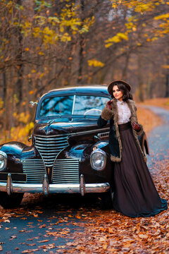 Beautiful female in vintage  dress, lace blouse and hat with veil standing near retro brown car on autumn background. Elegant lady in gloves posing on nature.Warm art work.