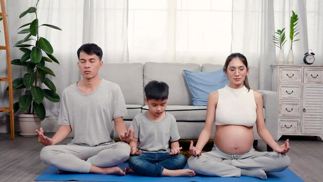 4K, Asian parent and son sitting in yoga meditation pose in living room. son follows father in yoga poses. Pregnant mother wearing a tank top reveals stomach, woman relaxing while pregnant