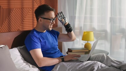 Young man with bionic prosthesis lying in bed and having video chat on laptop