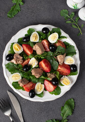 Salad with arugula, cod liver, quail eggs, olives and cherry tomatoes on white plate on dark gray background, Top view