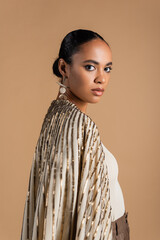 young african american model in golden jewelry looking at camera isolated on beige.