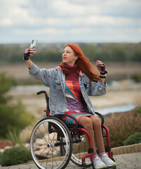 Beautiful red-haired girl on a wheelchair in a city park with a smartphone. Walks in the open air. Socialization of the disabled. Remote work. accessible environment.
