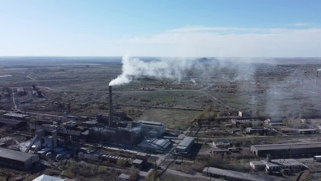 Puffs of white smoke are emitted from the chimneys against the blue sky. Old metallurgical plant. Industrial factory with many buildings. Environmental pollution. Drone flying next to the smoke