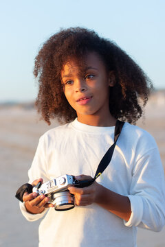 Portrait of thoughtful girl with old-fashioned camera. Female model with curly hair and in white pullover holding camera on beach. Childhood, hobby concept
