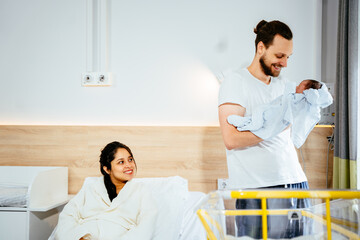 Happy caucasian man holding baby while wife resting in hospital bed after childbirth. Multiethnic couple with new born baby.