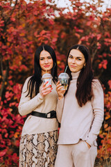 A beautiful young brunette woman and her friend, sister long-haired girl stand in nature in autumn with plastic cups with a drink and a straw. Photography, friendship.