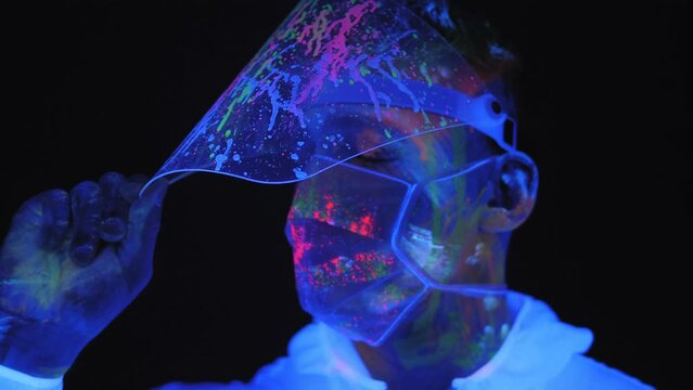 A girl in a white luminous protective suit and a protective mask in the dark under the light of fluorescent lamps, her hands and skin splattered with ultraviolet paint. Body art glows under UV light.