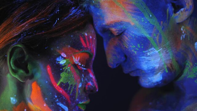 Couple with fluorescent makeup posing at camera, unusual creative colorful pattern on their skin. Attractive woman and man hug each other by the light of fluorescent lamps.