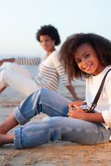 Smiling girl with old-fashioned camera. Female model with curly hair and in white pullover holding camera on beach. Mother in background. Childhood, hobby concept