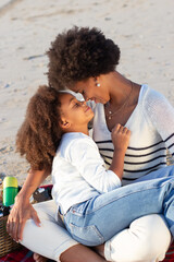 Caring African American family on picnic on beach. Mother and daughter in casual clothes sitting on blanket, hugging, eyes closed. Family, relaxation, nature concept