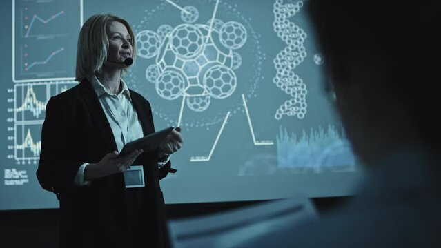 Selective focus of confident senior businesswoman wearing wireless microphone set holding digital tablet making presentation of new product