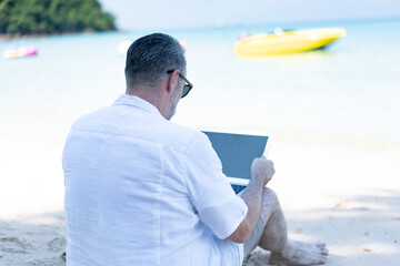 Senior man working on his laptop on the beach, Businessman who has a business meeting over a smartphone. Man sits on the beach with a laptop and examines the information. 