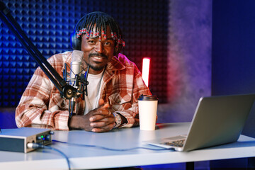 African radio host sitting at desk recording in studio with microphone and laptop