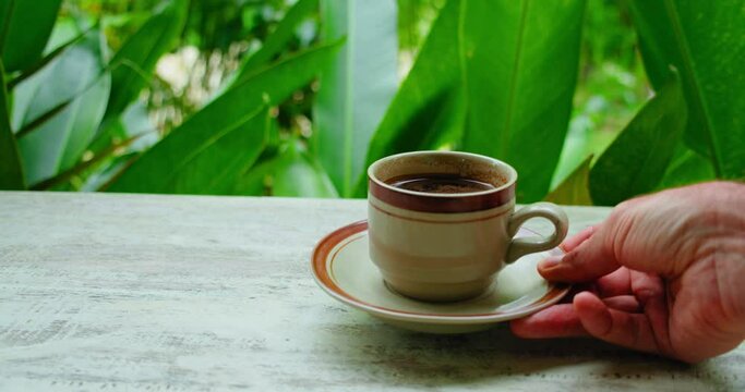 Table in a green lush tropical garden. Waiter serving a cup of black coffee on countertop. Male hand bringing a drink. Outdoor beverage made with espresso doppio americano lungo decaf.