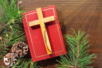 Wood cross laying on a closed red Christian bible with green fir branches and cones on a wooden...