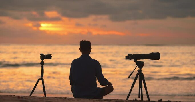 Man photographer with professional cameras long lens and tripods sitting next to his equipment meditating and waiting for it to record a timelapse 4K