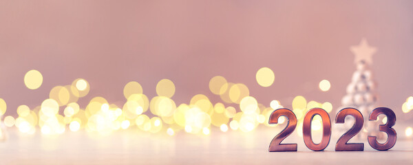 Metal numbers 2023 on a white table with Christmas trees and bokeh lights. Happy New Year 2022 is...