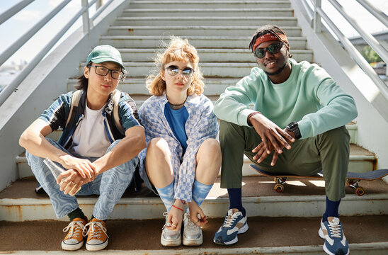 Diverse group of young people wearing street style clothes outdoors while sitting on stairs in urban area and looking at camera