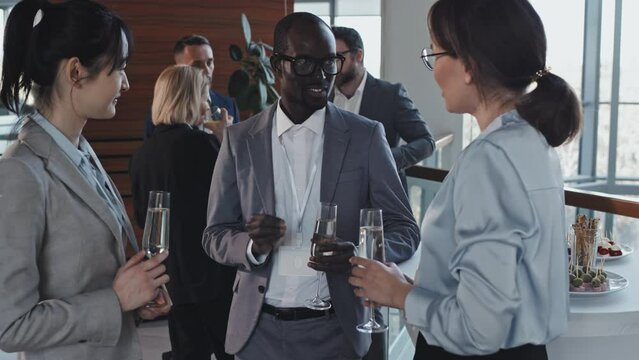 Handsome young Black man standing relaxed at buffet table telling funny story to his female Asian co-workers at business event party