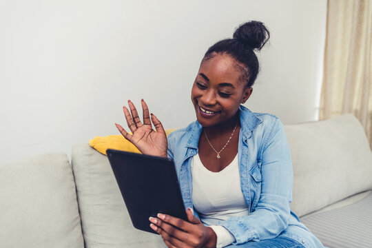 Smiling young African American woman sit on sofa at home wave greet talking speaking on video call on pad. Happy biracial female have webcam digital virtual event on tablet. Communication concept.