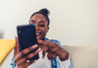 Close up of a young African woman using a phone at home. Shot of a young women wearing headphones to listen to music on her cellphone.