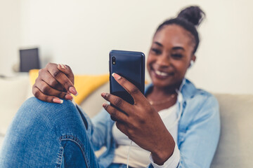 Close up of a young African woman using a phone at home. Shot of a young women wearing headphones to listen to music on her cellphone.