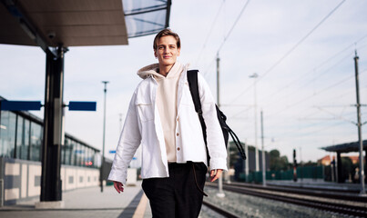 Woman wearing casual white clothes walking on railroad station platform with backpack.