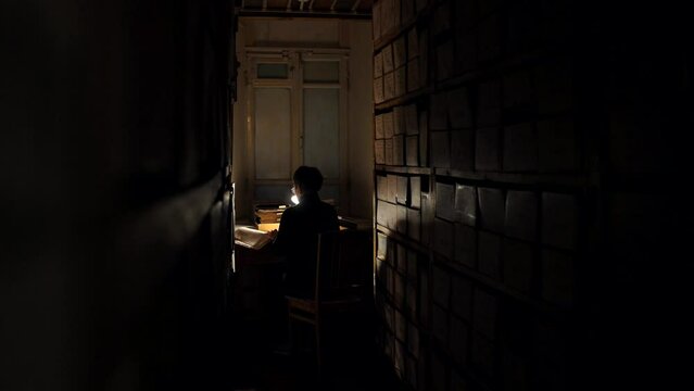 A woman reads historical documents from an old book in a dark library among rows of books by the light of a table lamp. An archivist and historian studies archival documents by turning over the pages.