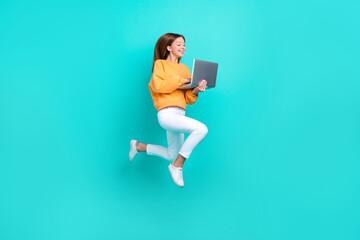 Full length photo of excited funny schoolgirl wear orange sweatshirt typing modern device jumping high isolated turquoise color background