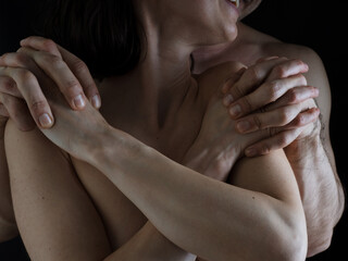 A man hugs a woman from behind. Naked bodies. Plexus of the hands. Close-up photo without a face on a dark background.