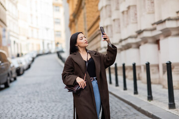 stylish brunette woman with crossbody taking selfie on cellphone in prague.